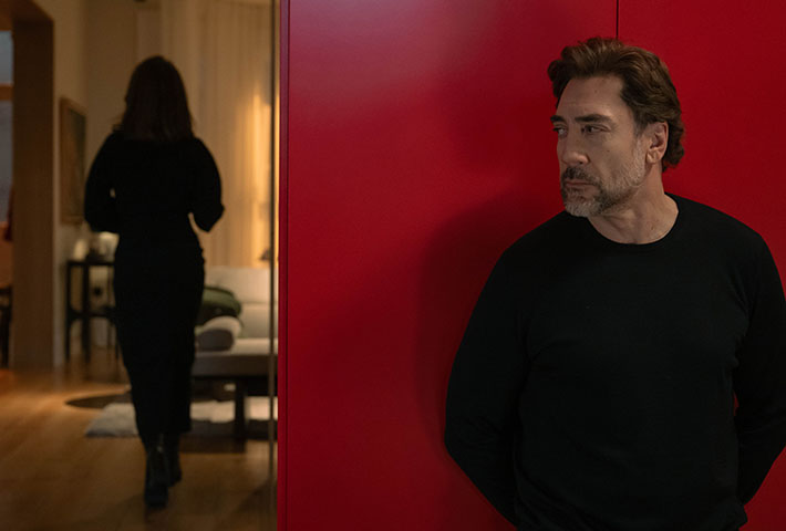 Javier Bardem adossé à un mur rouge dans Bunker, tandis qu'une femme vêtue de noir s'éloigne dans la pièce voisine