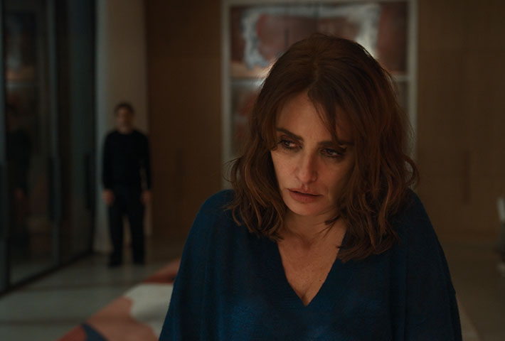 Penélope Cruz au premier plan dans Bunker, le regard baissé, avec une silhouette masculine en retrait dans le fond de la pièce